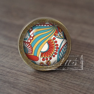 May include: A round, brass-coloured cabinet knob with a colourful, abstract design. The design features red, yellow, blue, and white colours.