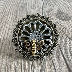 May include: A silver and gold metal brooch with a bee design in the center. The bee is surrounded by a silver flower with a cut-out design. The flower is surrounded by a circle of small silver flowers.