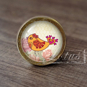 Può includere: Un pomello per armadio rotondo in ottone con un inserto in vetro che presenta un uccello giallo con accenti rosa e un design floreale. L'uccello ha una coda a forma di cuore rosa ed è circondato da rose rosa.