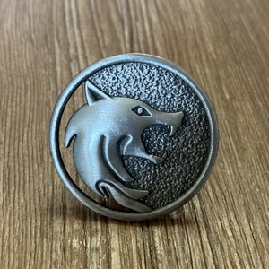 以下が含まれることがあります： 狼の頭部のデザインが施された銀色の金属製のピン。狼の頭部は横向きで、口が開いて歯が見える。ピンは円形で、表面にテクスチャがある。