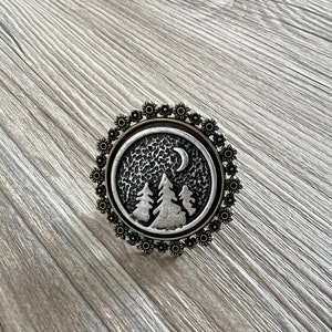 Puede incluir: Un botón de metal plateado con un diseño detallado de una luna creciente y tres pinos sobre un fondo texturizado. El botón está rodeado por un marco decorativo, ornamentado, de color bronce.