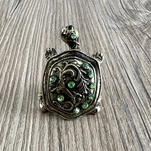 Op de afbeelding: Een gouden schildpadfiguur met groene strasssteentjes op zijn schild. De schildpad staat op een houten oppervlak.