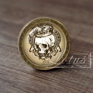 Puede incluir: Perilla de armario redonda de estilo antiguo con una calavera coronada. La calavera está en el centro, rodeada de detalles ornamentados. La perilla tiene un acabado dorado y está sobre una superficie de madera.