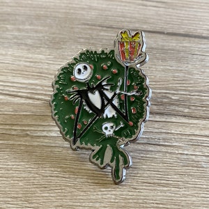 以下が含まれることがあります： ナイトメア・ビフォア・クリスマスのジャック・スケリントンが描かれた銀色の金属製ピン。彼はトレードマークの白黒の衣装を着て、赤と黄色のギフトボックスを持っています。彼は赤い実のついた緑のリースの前に立っています。