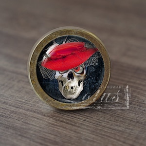 Pode incluir: Um botão de armário redondo de cor latão com um inserto de vidro que apresenta uma caveira com um chapéu vermelho e um véu preto. A caveira tem dentes brancos e olhos pretos.