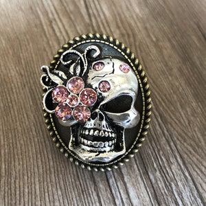 Pode incluir: Um crânio de prata com strass rosa engastados em um anel de cor bronze. O crânio tem um design floral de um lado e um rosto detalhado com uma boca preta.