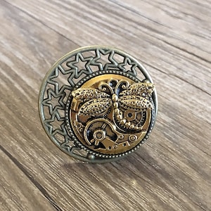 May include: A gold-toned metal ring with a decorative design featuring a dragonfly and gears. The ring is surrounded by a circle of stars.