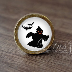 Pode incluir: Puxador de armário redondo com tema de Halloween. O puxador apresenta um fundo branco com um desenho de fantasma preto e morcegos. O fantasma tem um rosto laranja brilhante. O puxador tem uma borda de metal dourado.