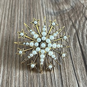 Peut inclure: Une broche en forme d'étoile dorée avec un motif d'étoile centrale, entourée de petites étoiles et ornée de perles blanches.
