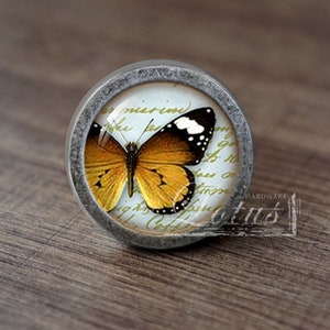 May include: A round, silver-toned cabinet knob with a glass insert featuring a yellow and black butterfly on a white background with handwritten text.