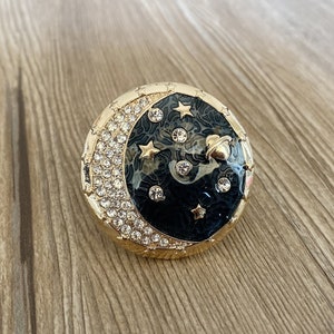 以下が含まれることがあります： 黒のエナメルで三日月模様が施されたゴールドトーンのブローチ。三日月はラインストーンで囲まれており、小さな金の星と惑星のデザインが施されています。