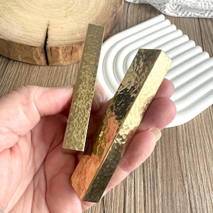 Puede incluir: Dos tiradores de armario rectangulares de latón martillado. Los tiradores tienen una superficie texturizada y un cálido tono dorado. Los tiradores de armario se sostienen en una mano, con una superficie de madera y un objeto decorativo blanco en el fondo.