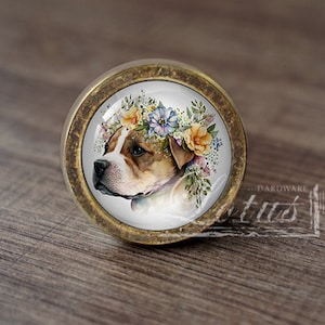 May include: A round, brass cabinet knob with a glass insert featuring a painted image of a brown and white dog wearing a flower crown. The dog is looking to the left.