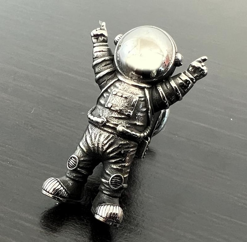 May include: A silver astronaut figurine with raised arms and a helmet. The astronaut is standing on a dark surface.