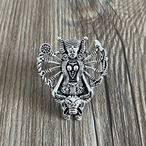 May include: A silver-colored pendant featuring a multi-armed deity figure. The pendant has intricate details, including a headdress, multiple arms, and a detailed face. The design is symmetrical and appears to be a religious or spiritual symbol.