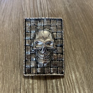 May include: A silver skull ring with a woven design. The skull is detailed with eye sockets, a nose, and a mouth.