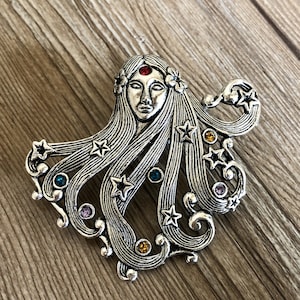 Può includere: Un broche argentato con una donna stilizzata con lunghi capelli fluenti, adornato con strass colorati. I capelli sono rappresentati come ciocche fluttuanti con stelle e volute, creando un design fantasioso.