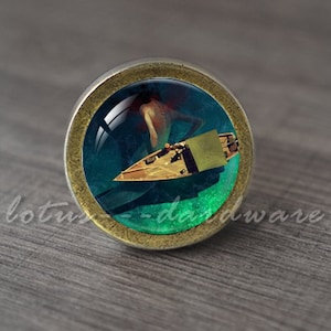 May include: A round, antique-style cabinet knob with a glass insert featuring a surreal image of a figure in water near a boat. The knob has a bronze-colored metal base and a dark teal and green background.
