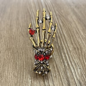 K&ouml;nnte beinhalten: Ein goldener Skeletthandring mit roten und klaren Strasssteinen. Der Ring ist mit einem Totenkopf- und Knochenkreuz-Design verziert.
