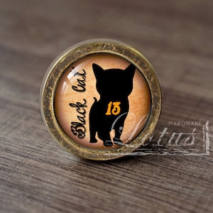 Może przedstawiać: Okrągła gałka do szafki z antycznego mosiądzu z czarną sylwetką kota i numerem 13 w kolorze złotym. Napis "Black Cat" jest napisany kursywą. Gałka ma jasnobrązowe tło.