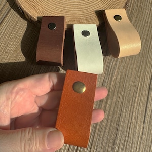 Könnte beinhalten: Eine Auswahl an Leder-Schubladengriffen in verschiedenen Farben: Braun, Weiß, Orange und Beige. Jeder Griff hat eine rechteckige Basis, einen gebogenen Griff und einen Messingniet. Präsentation auf einer Holzoberfläche.
