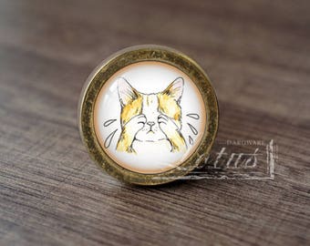 May include: A round, gold-coloured cabinet knob with a white, round, glass insert. The glass insert features a cartoon illustration of a cat with its paws covering its eyes, as if crying.