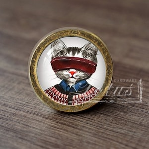 Op de afbeelding: Een ronde, messingkleurige kastknop met een witte achtergrond en een cartoonafbeelding van een kat met een rood blinddoek en een blauw-rode trui.