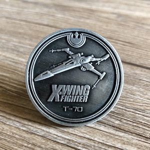 May include: A silver-colored metal coin with a black background featuring a detailed image of an X-Wing fighter from Star Wars. The coin also includes the text "X-WING FIGHTER" and "T-70".