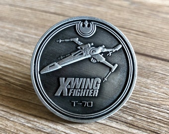 Può includere: Una moneta di metallo color argento con sfondo nero che presenta un'immagine dettagliata di un caccia X-Wing di Star Wars. La moneta include anche il testo "X-WING FIGHTER" e "T-70".