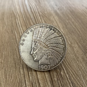 Puede incluir: Una moneda de plata con el perfil de una mujer nativa americana con un tocado de plumas. La moneda tiene el año 1907 inscrito en ella.
