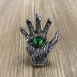 Op de afbeelding: Een zilveren hand met een groen oog in de palm. De hand is gedetailleerd met getextureerde lijnen en een verhoogd oog.
