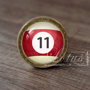 May include: A brass cabinet knob with a red, white, and cream colored billiard ball design. The ball has the number "11" printed in black.