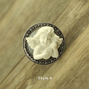 May include: A silver-toned brooch with a white cherub playing a musical instrument. The cherub is surrounded by a decorative border with a black background.