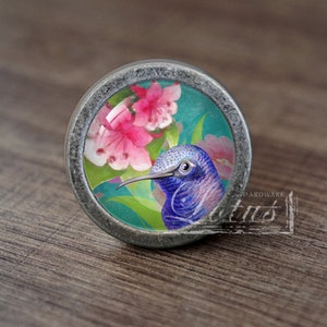 Peut inclure: Un bouton argenté avec un insert en verre rond représentant un colibri violet avec des fleurs roses et des feuilles vertes.