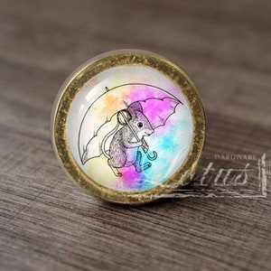 以下が含まれることがあります： 真鍮仕上げの丸いキャビネットノブ。カラフルな水彩画の背景に、傘を持ったネズミの気まぐれなイラストが特徴です。画像は透明なドーム型のガラスに収められています。