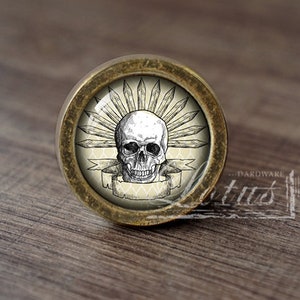 Puede incluir: Un pomo de armario redondo de color latón con un diseño de calavera. La calavera está rodeada por un anillo de púas y una pancarta con un patrón decorativo. El pomo está sobre una superficie de madera.