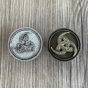 Op de afbeelding: Twee ronde metalen knoppen met een Keltisch knoopontwerp in de vorm van een triskelion. De knop links is zilver en de knop rechts is goud.