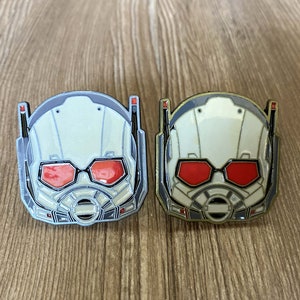 May include: Two Ant-Man character enamel pins. The pins are silver and gold in color and feature a white helmet with red eyes and a black mouth.