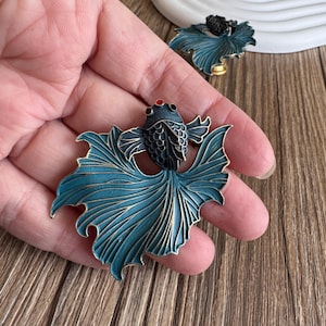 Könnte beinhalten: Ein Paar teal-farbene und schwarze Fischbroschen. Die Broschen haben detaillierte Flossen und Schuppen mit roten Akzenten auf den Augen. Die Fische werden in einer Hand gehalten, mit einer Holzoberfläche im Hintergrund.