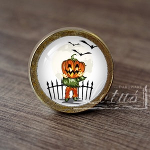 Pode incluir: Um botão de armário redondo de cor bronze com um inserto de cerâmica branca que apresenta uma abóbora de desenho animado com rosto, vestindo uma camisa verde e de pé em frente a uma cerca preta. Os morcegos voam no fundo contra uma lua branca.