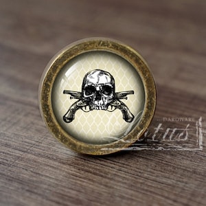 Könnte beinhalten: Ein runder, messingfarbener Schrankknopf mit einem Glaseinsatz, der ein schwarz-weißes Totenkopf- und Knochenkreuzdesign zeigt. Der Totenkopf ist von einem weißen und beigefarbenen gemusterten Hintergrund umgeben.