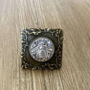 May include: A square-shaped metal button with a floral design around the edges. The center of the button features a silver-toned bee with a detailed design.