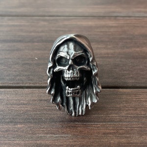 Op de afbeelding: Een zilverkleurige schedelring met een kapontwerp. De schedel heeft een open mond en gedetailleerde gelaatstrekken. De ring is gemaakt van metaal en is waarschijnlijk een accessoire in gotische of biker-stijl.