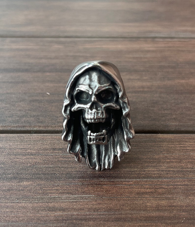 Op de afbeelding: Een zilverkleurige schedelring met een kapontwerp. De schedel heeft een open mond en gedetailleerde gelaatstrekken. De ring is gemaakt van metaal en is waarschijnlijk een accessoire in gotische of biker-stijl.