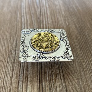 May include: A silver square drawer pull with a raised floral design. The center of the pull features a gold bee with a textured surface.