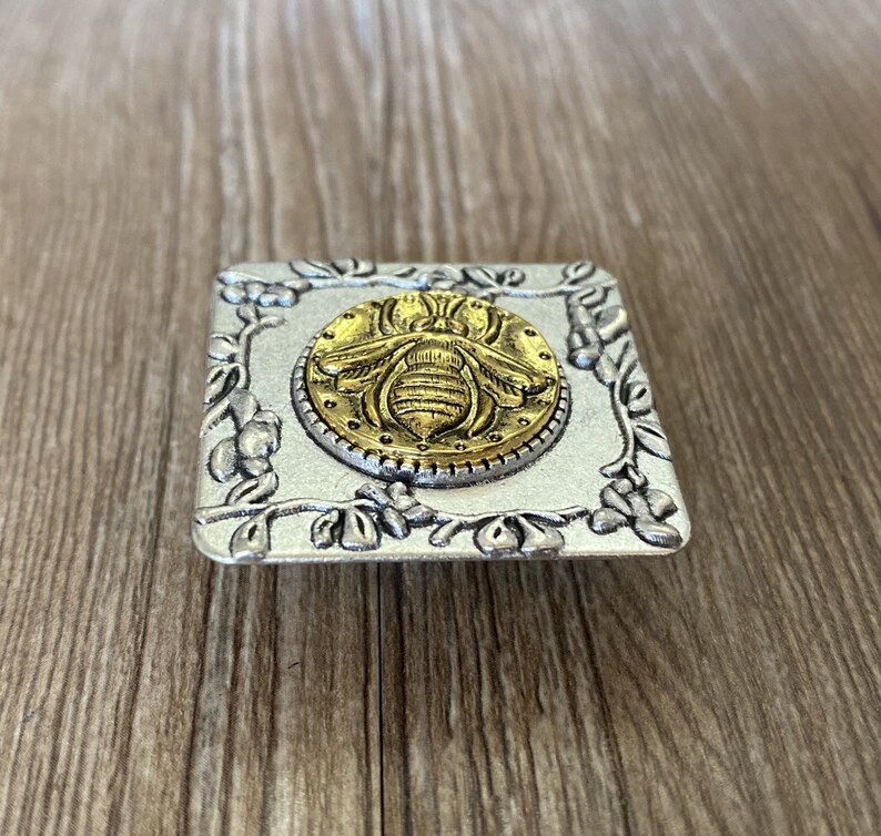May include: A silver square drawer pull with a raised floral design. The center of the pull features a gold bee with a textured surface.
