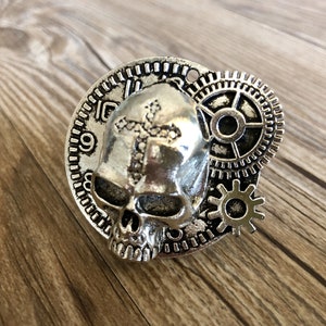 May include: Silver skull pendant with a cross design and gears. The skull has a detailed face and is surrounded by a clock face and gears.