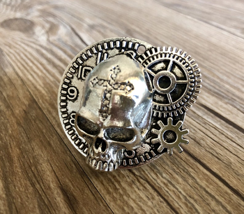 May include: Silver skull pendant with a cross design and gears. The skull has a detailed face and is surrounded by a clock face and gears.