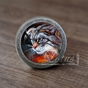 May include: A round, silver-toned cabinet knob with a glass insert featuring a close-up image of a wildcat with green eyes. The cat is looking to the left of the image.