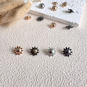 May include: A collection of sun-shaped metal embellishments in various metallic finishes, including gold, silver, and dark bronze. These decorative elements are arranged on a textured, off-white surface, suggesting a craft or jewelry-making theme.